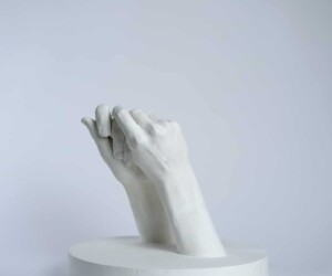 A white ceramic sculpture of two cast hands atop a pedestal against a white background