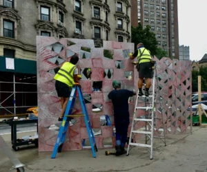 A photo of three workers examining a sculpture. Two of the workers are wearing yellow safety vests and are positioned on ladders. The third worker is standing beneath them on the ground.