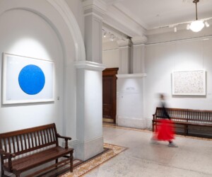 A photograph of a hallway with wooden benches. On the walls are framed prints. There is a person walking through the space who is blurred and they are wearing a red skirt.