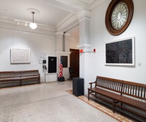 A photograph of a hallway with wooden benches. On the walls are framed prints.