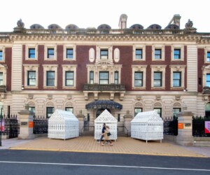 A photograph of people walking in front of three white, ornate sculptural houses outside the Cooper Hewitt Museum.