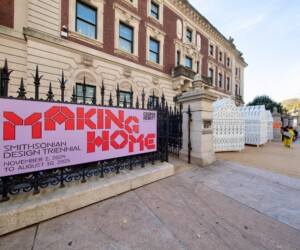A photograph of a sign that reads "Making Home" Smithsonian Design Triennial November 2, 2024 to August 10, 2025. In the background are three white, ornate sculptural houses with people walking in front of them.