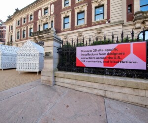 A photograph of a sign that reads "Discover 25 site-specific installations from designers and artists across the U.S., U.S. territories, and Tribal Nations." In the background are three white, ornate sculptural houses.